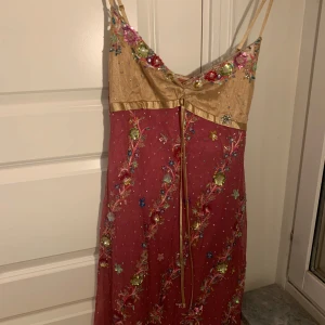 Rosa och guld klänning från Galliano - Elegant rosa klänning med guld detaljer från Galliano. Klänningen har tunna axelband och är dekorerad med färgglada broderier och pärlor. Den har en vacker knytning framtill och en unik design som verkligen sticker ut. Material är silke, polyester och bomull