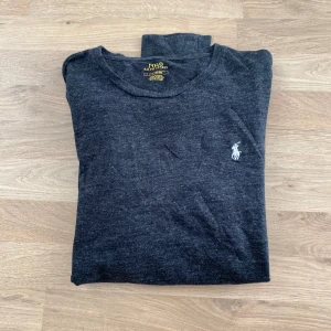 Polo Ralph Lauren Långärmad Tröja - Säljer en stilren mörkgrå tröja från Polo Ralph Lauren med det klassiska broderade logotypen på bröstet. Tröjan har långa ärmar och en rund halsringning. Perfekt för en avslappnad och snygg look.