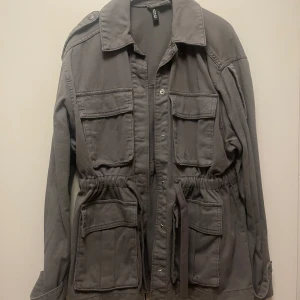 Grå jacka från H&M - Säljer en grå jacka från H&M med knappar och knytband i midjan. Jackan har flera fickor framtill och långa ärmar. Perfekt för en stilren look. Storlek xs men ganska oversize så passar mig som är S. Endast använd vid ett tillfälle, nästintill oanvänd.