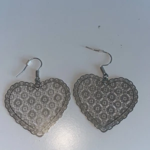 Silvriga hjärtformade örhängen - Snygga silvriga örhängen i form av hjärtan med ett intrikat mönster. Perfekta för att ge en romantisk touch till din outfit. De är lätta och bekväma att bära hela dagen.