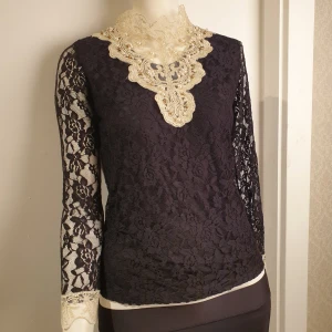 Svart spetsblus med pärldetaljer - Elegant svart spetsblus med långa ärmar och vacker beige spetskrage dekorerad med pärlor. Blusen har ett intrikat blommönster och en stilren passform. Perfekt för speciella tillfällen.
