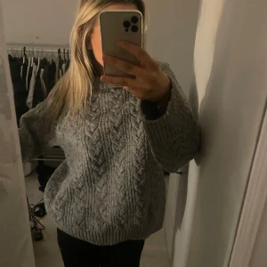 Grå stickad tröja från H&M - Mysig grå stickad tröja från H&M med ett klassiskt flätmönster. Perfekt för kyliga dagar med sin varma och bekväma design. Tröjan har långa ärmar och ribbade kanter för extra komfort. Ett måste i garderoben för höst och vinter!
