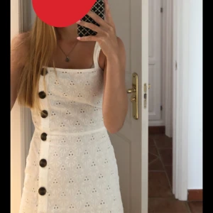 Vit klänning - Säljer en söt vit klänning med broderade detaljer och stora knappar framtill. Klänningen har smala axelband och en kort längd. Normal i storlek! 