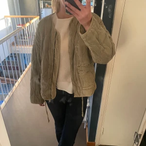 Olivgrön quiltad jacka - Säljer en olivgrön quiltad jacka med dragkedja och snörning i nederkant. Jackan har långa ärmar och två praktiska fickor framtill. Perfekt för kyligare dagar.