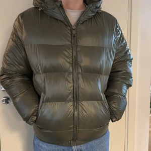 Olivgrön Zara pufferjacka med huva - Snygg olivgrön pufferjacka med dragkedja och huva. Jackan har en glansig yta och är perfekt för kyligare dagar. Den har långa ärmar och en bekväm passform som håller dig varm och stilren.