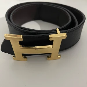 Svart bälte med H-spänne - Snyggt svart bälte med ett stort H-format spänne i guldfärg. Perfekt för att ge en stilren touch till din outfit. Bältet är tillverkad i skinn och har en klassisk design som passar till många olika stilar.  Den passar även bra till både jeans och kostymbyxor. Vid funderingar så äre bara o fråga. Pris kan diskuteras..