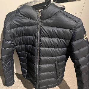 Mörkblå dunjacka från Colmar - Säljer en snygg mörkblå dunjacka från Colmar. Jackan har en dragkedja framtill och långa ärmar. Perfekt för kyligare dagar med sin pufferdesign som håller värmen. Den har en stilren look med en liten logga på ärmen.