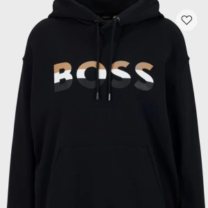 Svart hoodie från Boss - Snygg svart hoodie från Boss med stor logga i vitt och guld på bröstet. Den har en klassisk känguruficka och justerbar dragsko i huvan. Perfekt för en avslappnad stil. Den är 100 procent äkta och används ett fåtal gånger. Priset kan diskuteras. Obs bilden är inte hur tröjan ser ut idag efter användning vill man ha ytterligare kan man kontakta mig. 
