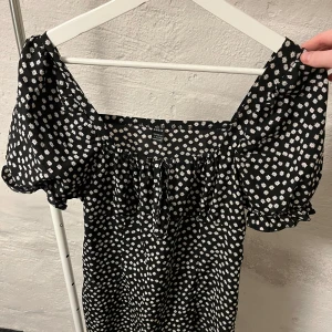 Svart blommig klänning - Charmig svart klänning med vita blommor och korta puffärmar. Man kan styra justera vid bysten. Perfekt för en somrig look!