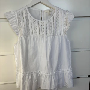 Vit blus med spetsdetaljer från H&M - Säljer en söt vit blus från H&M med spetsdetaljer på framsidan och volangärmar. Blusen har en rund halsringning och en lätt volang nertill. Aldrig använd🤍