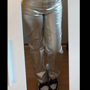 Silvriga byxor - Snygga silvriga byxor med hög midja och rak passform. Perfekta för att sticka ut med en glittrig look. Byxorna har en dragkedja framtill och är tillverkade i ett syntetmaterial som ger en glänsande finish.