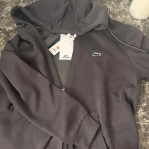 Svart hoodie från Lacoste - Säljer en stilren svart hoodie från Lacoste med dragkedja och den ikoniska krokodilloggan på bröstet. Hoodien har långa ärmar och en praktisk huva med dragsko. Perfekt för en avslappnad stil.
