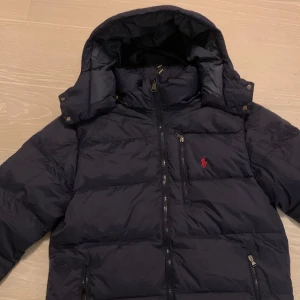Mörkblå dunjacka från Polo Ralph Lauren - Säljer en snygg mörkblå dunjacka från Polo Ralph Lauren med logga på bröstet. Jackan har dragkedja och en avtagningsbar luva som går att stänga med knappar eller. Perfekt för kyliga dagar. Det är medium och är normal i storleken.