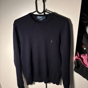  Polo Ralph Lauren - Säljer en stilren mörkblå tröja från Polo Ralph Lauren i slim fit. Tröjan har en liten grön logga på bröstet och är tillverkad i ett mjukt material. Perfekt för en klassisk look.
