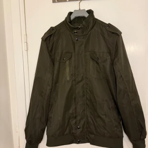 olivgrön jacka - Snygg olivgrön jacka med knappar och dragkedja. Den har långa ärmar och ribbade muddar vid ärmslut och nederkant. Perfekt för höst och vår med en stilren look.