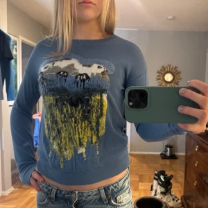 Blå tröja med konstnärligt motiv - Säljer en blå tröja med ett unikt konstnärligt motiv i flera färger på framsidan. Tröjan har långa ärmar och en normal passform. Perfekt för dig som vill ha något annorlunda i garderoben!