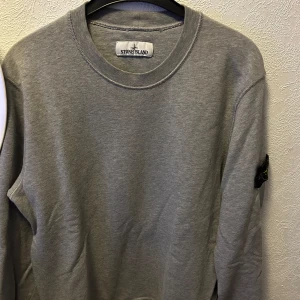 Grå tröja från Stone Island - Säljer en stilren grå tröja från Stone Island. Tröjan har långa ärmar och en rund halsringning. Perfekt för en avslappnad stil med den ikoniska loggan på ärmen.