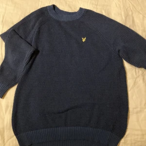Blå stickad tröja från Lyle & Scott - Säljer en snygg blå stickad tröja från Lyle & Scott med deras ikoniska logga på bröstet. Tröjan har en ribbad krage och långa ärmar, perfekt för kyligare dagar. Passar bra till jeans för en avslappnad stil.