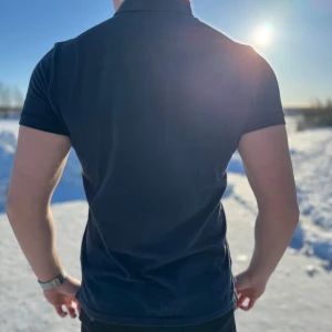 Mörkblå pikétröja från Ralph Lauren - Snygg mörkblå pikétröja från Ralph Lauren med klassisk krage och knappdetaljer. Perfekt för en stilren look. Den har en liten broderad logga på bröstet.