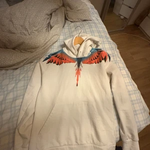 Vit hoodie med färgglatt vingmönster från Marcelo Burlon - Säljer en vit hoodie från Marcelo Burlon med ett iögonfallande vingmönster i rött, blått och svart över axlarna. Hoodien har en klassisk passform med långa ärmar och en bekväm huva. Perfekt för att ge din outfit en unik touch!