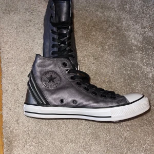  Converse All Star  - Snygga gråa  Converse All Star i skinn med coola dragkedjor på baksidan. Klassisk design med snörning och vit sula. Perfekta för en stilren look med en twist.