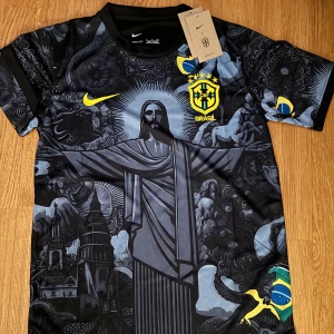 Svart Brasilien fotbollströja från Nike - Snygg svart fotbollströja från Nike med Brasilien-tema. Tröjan har ett unikt mönster med Kristusstatyn och andra detaljer i grått och blått. Den har korta ärmar och det ikoniska Brasilien-emblemet i gult på bröstet. Perfekt för fotbollsfans som vill sticka ut!