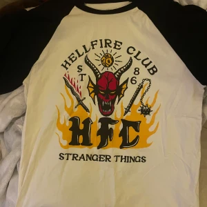 Hellfire Club Stranger Things T-shirt från Pull&Bear - Vit och svart t-shirt från Pull&Bear med Hellfire Club från Stranger Things-tryck. Tröjan har vit kropp och svarta ärmar, på framsidan finns ett stort tryck med Hellfire Clubs märket. Tröjan är köpt från Pull&Bear i storlek M och är endast använd ett fåtal gånger!