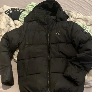 Svart pufferjacka från Calvin Klein Jeans - Säljer en stilren svart pufferjacka från Calvin Klein Jeans. Jackan har en dragkedja framtill och en broderad logga på bröstet samt en patch på ärmen. Perfekt för kyliga dagar med sin vadderade design och huva.
