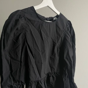 Svart blus med rosetter i rygg - Snygg svart blus från H&M med långa ärmar och  söta rosetter i ryggen. Perfekt för en stilren look. Blusen har en rund halsringning och är tillverkad i ett lätt material. Volang med resår nedtill och i ärmen nertill. Använd fåtal gånger!