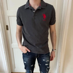 Polo Assn Piké  - Snygg mörkgrå pikétröja från USPA med en stor röd logga på bröstet.🇺🇸🇺🇸🐎