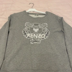 Grå sweatshirt från Kenzo - Säljer en grå sweatshirt från Kenzo med ett stort tigerhuvud och Kenzo-logga broderat på framsidan. Tröjan har långa ärmar och en rund halsringning. Perfekt för en avslappnad stil. Pris går att diskutera via köp! 