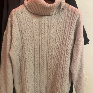 Beige stickad polotröja - Säljer en mysig beige stickad polotröja med ett klassiskt flätmönster. Tröjan har en hög polokrage och långa ärmar, perfekt för kyliga dagar. Den är både stilren och bekväm.