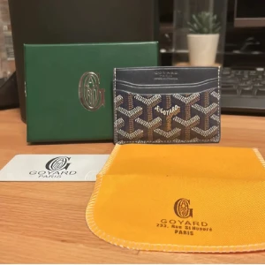 Korthållare  - Säljer en elegant plånbok med ett ikoniskt mönster i blått och beige. Plånboken är kompakt och perfekt för att hålla dina kort och kontanter organiserade. Den kommer med en snygg förvaringspåse i gult.