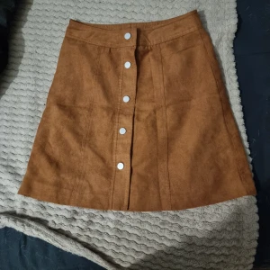 Brun mockakjol med knappar - Snygg brun mockakjol med knappar framtill. Kjolen har en A-linjeform och är perfekt för höstgarderoben. Den har en stilren design som gör den lätt att matcha med olika toppar. Aldrig använd bara provat en gång.