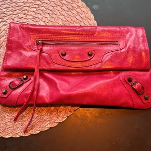 Cerise kuvertväska med dragkedja - Snygg cerise kuvertväska i skinn med dragkedja och dekorativa detaljer. Perfekt för att ge en färgklick till din outfit. Väskan har en praktisk storlek för att bära dina nödvändigheter.