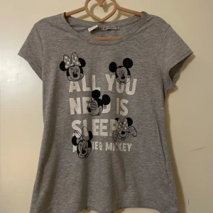 Grå Disney t-shirt  - Säljer en grå t-shirt från Disney med tryck av Musse och Mimmi samt texten 'All you need is sleep'. T-shirten har en rund hals och korta ärmar, perfekt för alla Disney-fans!