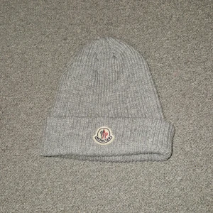 Grå stickad mössa från Moncler - Säljer en stilren grå stickad mössa från Moncler. Mössan har en uppvikt kant och ett broderat Moncler-märke framtill. Perfekt för kyliga dagar.
