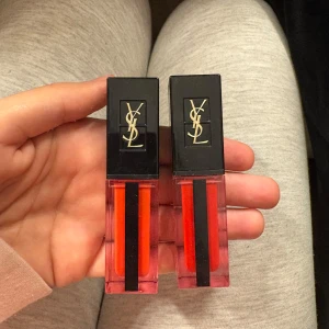 Oanvända Läppglans från YSL - Två eleganta läppglans från YSL i klara röda nyanser. Förpackningarna har en stilren design med svart lock och YSL-logotyp i guld. Perfekt för att ge läpparna en glansig och färgstark look.