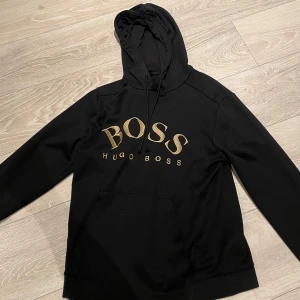 Svart hoodie från Hugo Boss - Säljer en stilren svart hoodie från Hugo Boss med guldtryck på bröstet. Den har en klassisk design med huva och dragsko samt en praktisk magficka. Perfekt för en avslappnad look.