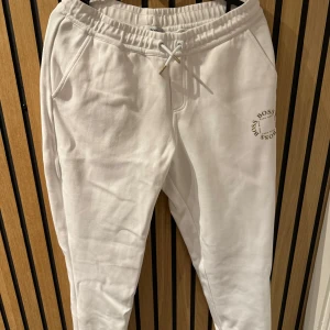 Vita mjukisbyxor från Hugo Boss - Snygga vita mjukisbyxor från Hugo Boss med elastisk midja och snörning. Byxorna har sidofickor och en bakficka, perfekt för en avslappnad stil. Boss-logga tryckt på sidan ger en extra touch av stil. Guld och vitt. Använda max 1 gång så ny skick. 16 år/174 cm från kidsbrandstore 