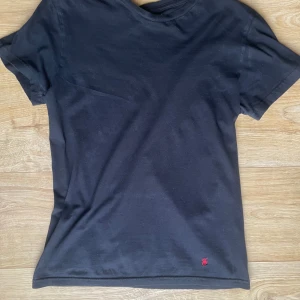 Svart t-shirt från Polo Ralph Lauren - Säljer en klassisk svart t-shirt från Polo Ralph Lauren med det ikoniska röda broderade logotypen på framsidan. T-shirten är kortärmad och tillverkad i mjuk bomull för en bekväm passform. Perfekt för en stilren look.