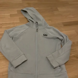 Grå hoodie från Under Armour - Säljer en grå hoodie från Under Armour med dragkedja och huva. Tröjan har en lös passform och är tillverkad i mjukt material, perfekt för kyliga dagar. Den har långa ärmar och en liten logotyp på bröstet.