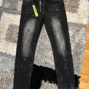 Svarta jeans med slitningar från Dsquared2 - Snygga svarta jeans från Dsquared2 med coola slitningar och en unik dragkedjedetalj. Jeansen har en modern slim passform och är perfekta för en trendig look. De har också en iögonfallande grön etikett som ger extra stilpoäng.