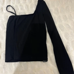 Svart oneshoulder topp från NA-KD - Snygg svart oneshoulder topp från NA-KD. Perfekt för en stilren look med en unik design som har en lång ärm och en axelbandslös sida. Passar utmärkt till både jeans och kjol.