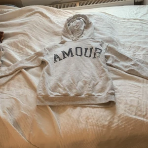 Grå hoodie från Gina Tricot med tryck - Säljer en grå hoodie från Gina Tricot med texten 'AMOUR' på framsidan och en stjärna på baksidan. Tröjan har långa ärmar och en bekväm huva. Perfekt för en avslappnad stil.Går ofta att sänka priset!