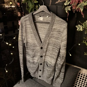 Grå kofta från Bläck - Säljer en stilren grå kofta från Bläck i storlek M. Koftan har en v-ringad design med knappar framtill och ribbade kanter. Perfekt för kyligare dagar och passar till många olika outfits.