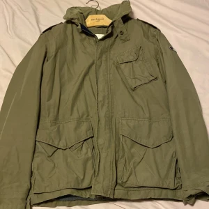 Field jacka   - Säljer en grön jacka från NN.07 i bra skick. Den har en dragkedja och knappar framtill, samt flera praktiska fickor. Perfekt för höst och vår med sin stilrena design och bekväma passform. Insidan är fodrad med mjukt material för extra komfort. Har luva också som man kan väckla in i jackan. Pris är inte hugget i sten