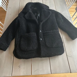 Svart teddyjacka från Monki - Mysig svart teddyjacka från Monki med stora fickor och knappar framtill. Perfekt för kyliga dagar. Jackan har en klassisk krage och är helfodrad för extra komfort. Pris kan diskuteras.
