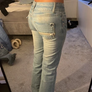 Ljusblå bootcut jeans - Snygga ljusblå jeans med låg midja och raka ben. Byxorna har unika detaljer som dubbla knappar och dekorativa fickor med dragkedja och broderi. Perfekta för en avslappnad stil.