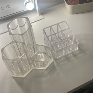 Transparenta sminkorganisatörer - Säljer två transparenta sminkorganisatörer i akryl. Den ena har tre fack i olika storlekar och den andra har nio mindre fack, perfekt för att organisera sminkborstar, läppstift eller andra små sminkprodukter.
